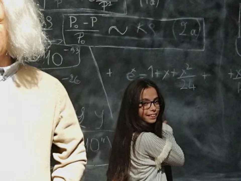 Two children stand by a chalkboard covered with equations, next to a wax figure resembling Albert Einstein in a classroom setting.