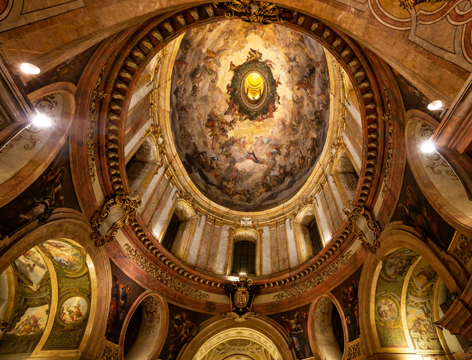Ornate domed ceiling with intricate frescoes, gold accents, arched windows, and a central medallion depicting a radiant face.