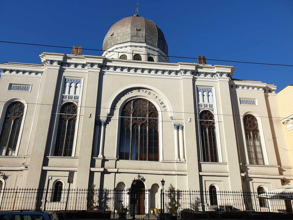 A large, ornate building with a dome, arched windows, and decorative elements. It has a fenced front and clear blue sky above.
