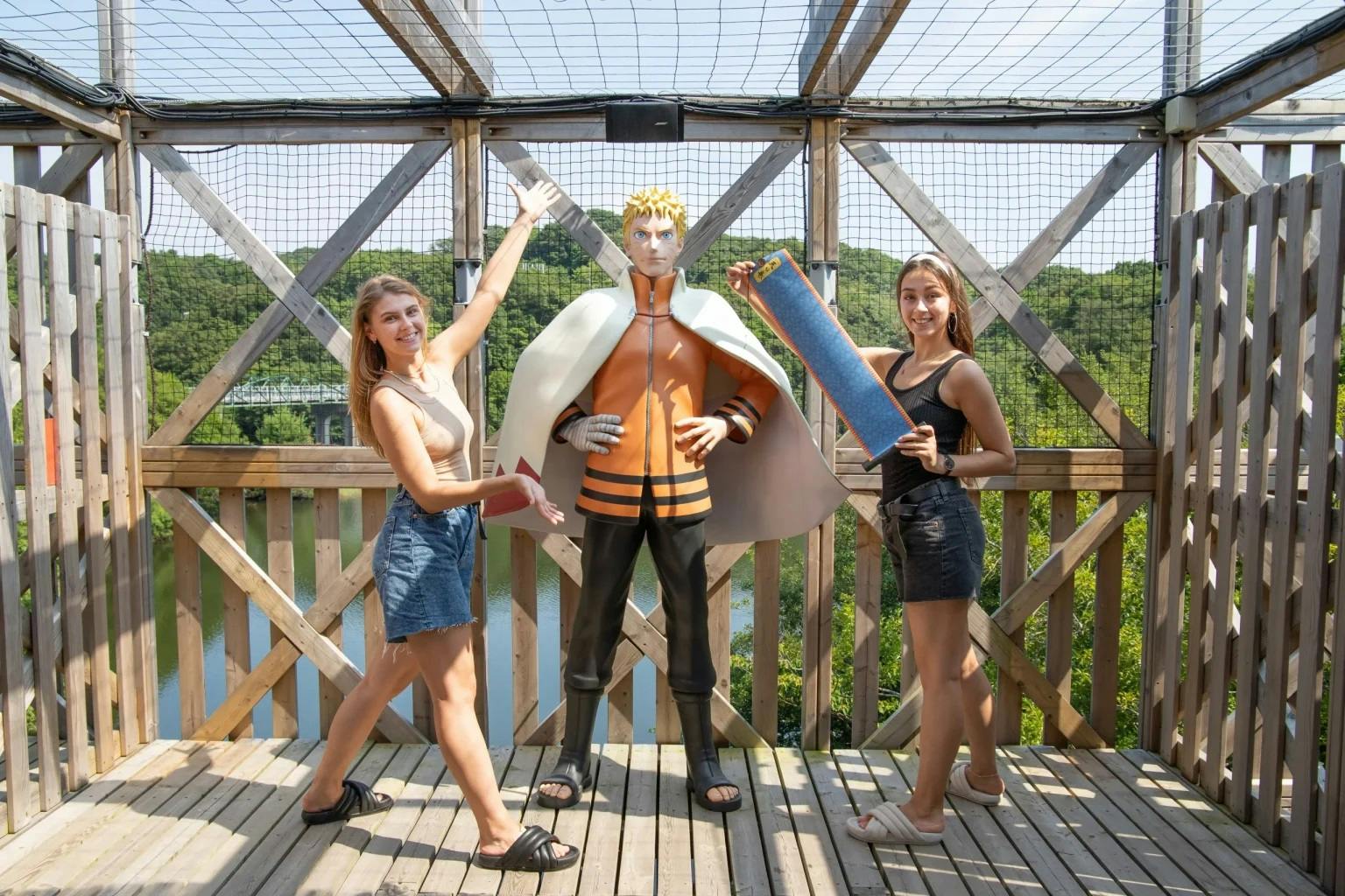 Two people pose with a tall statue of a character wearing a white cape and orange outfit, on a wooden platform outdoors.