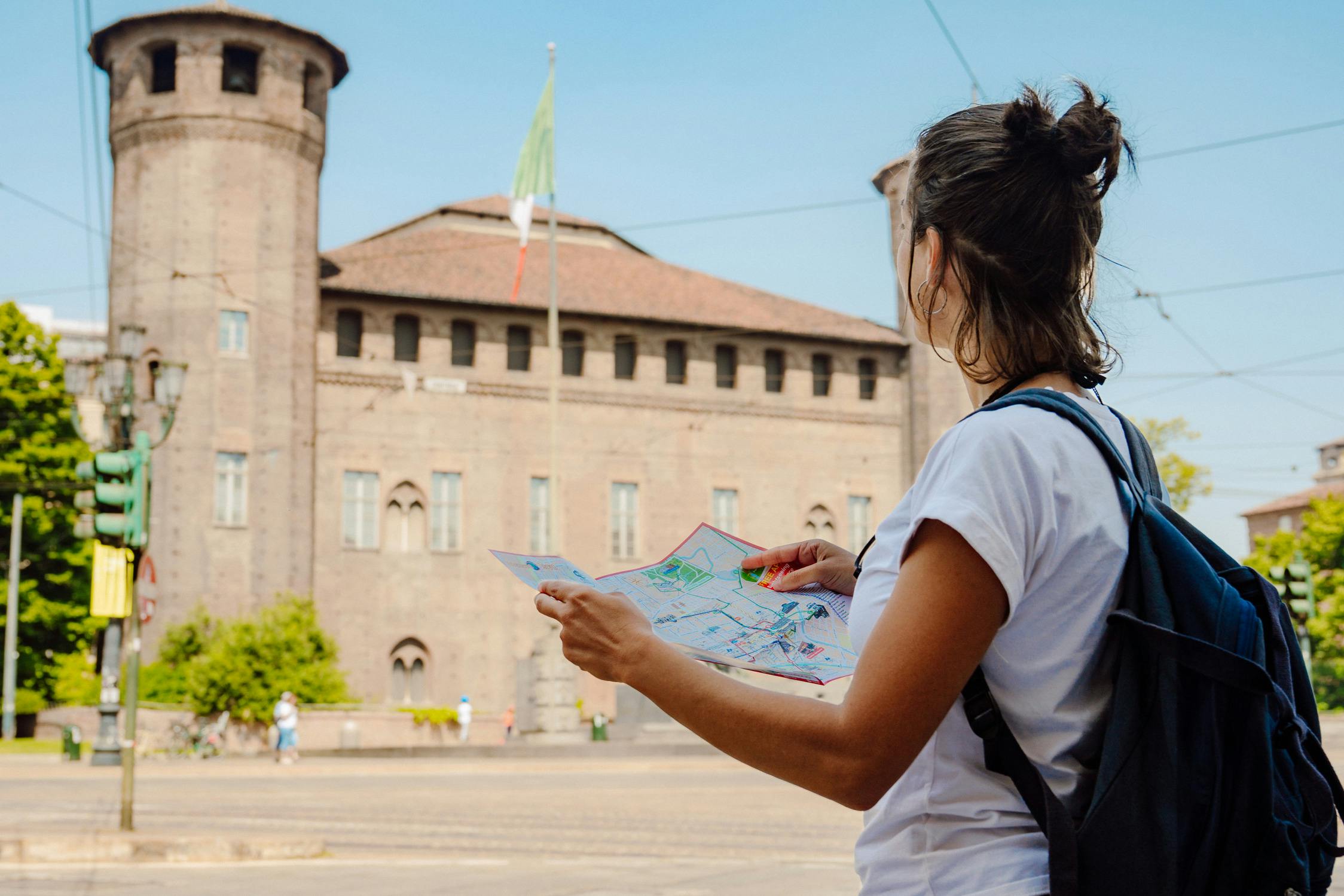 A person holding a map, standing in front of an old castle-like building with towers and a flag on a pole.