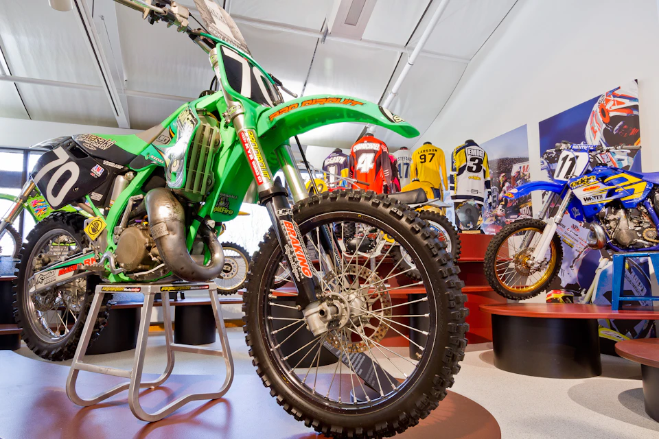 Close-up of a green dirt bike on display in a showroom, with motocross jerseys and another bike in the background.