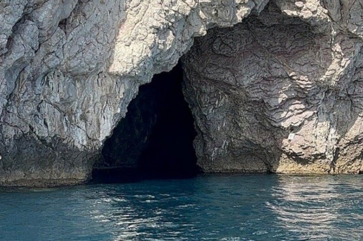 A rocky cave opening along a coastal cliff with blue water in the foreground.