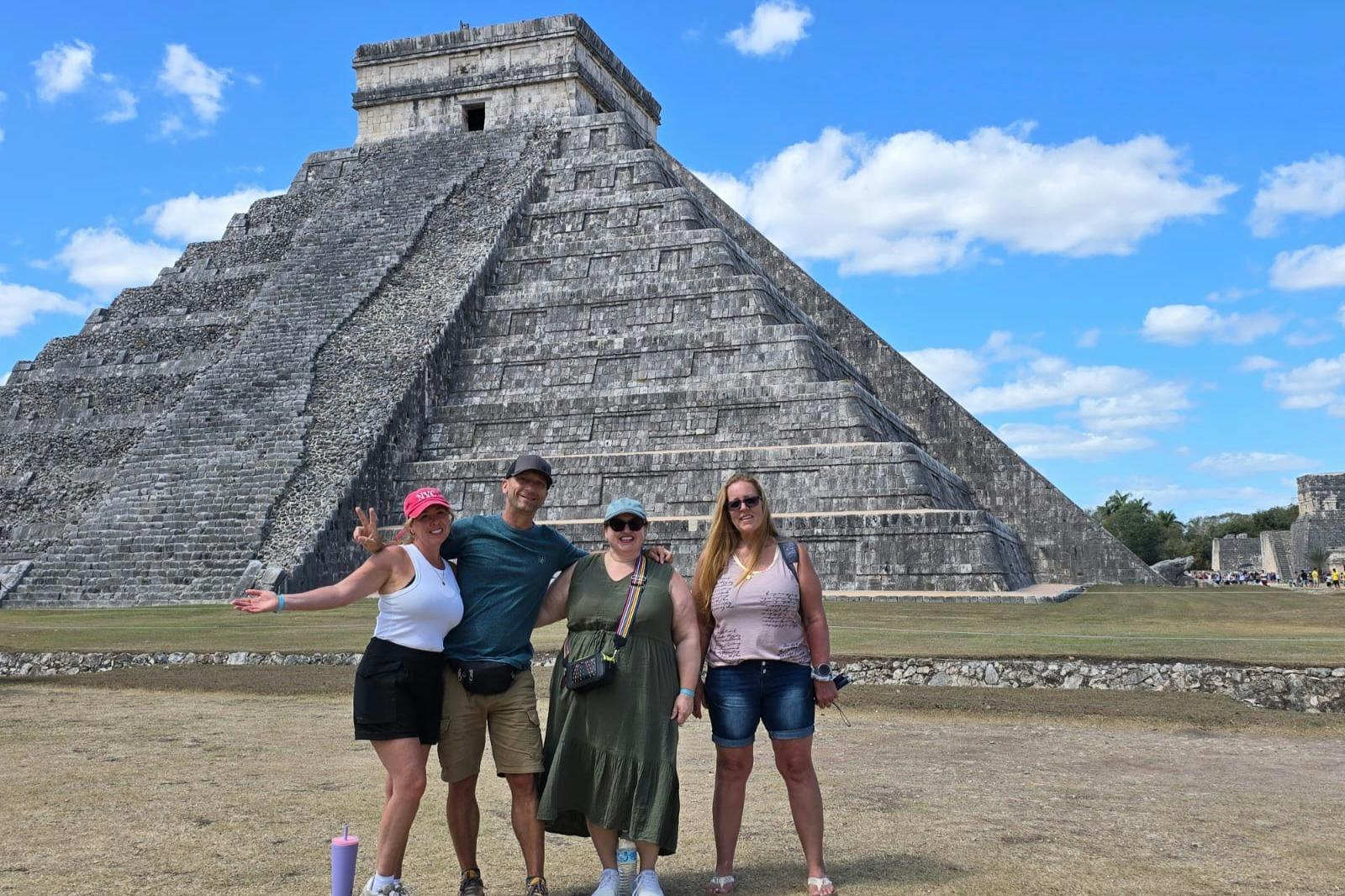 Четыре человека позируют перед большой каменной пирамидой под солнечным небом.