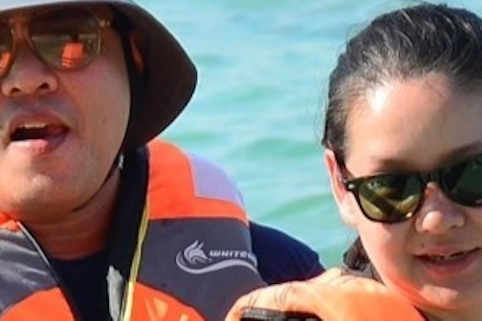 Two people in life jackets paddle a clear kayak on calm, blue water. Both wear sunglasses; one has a hat.