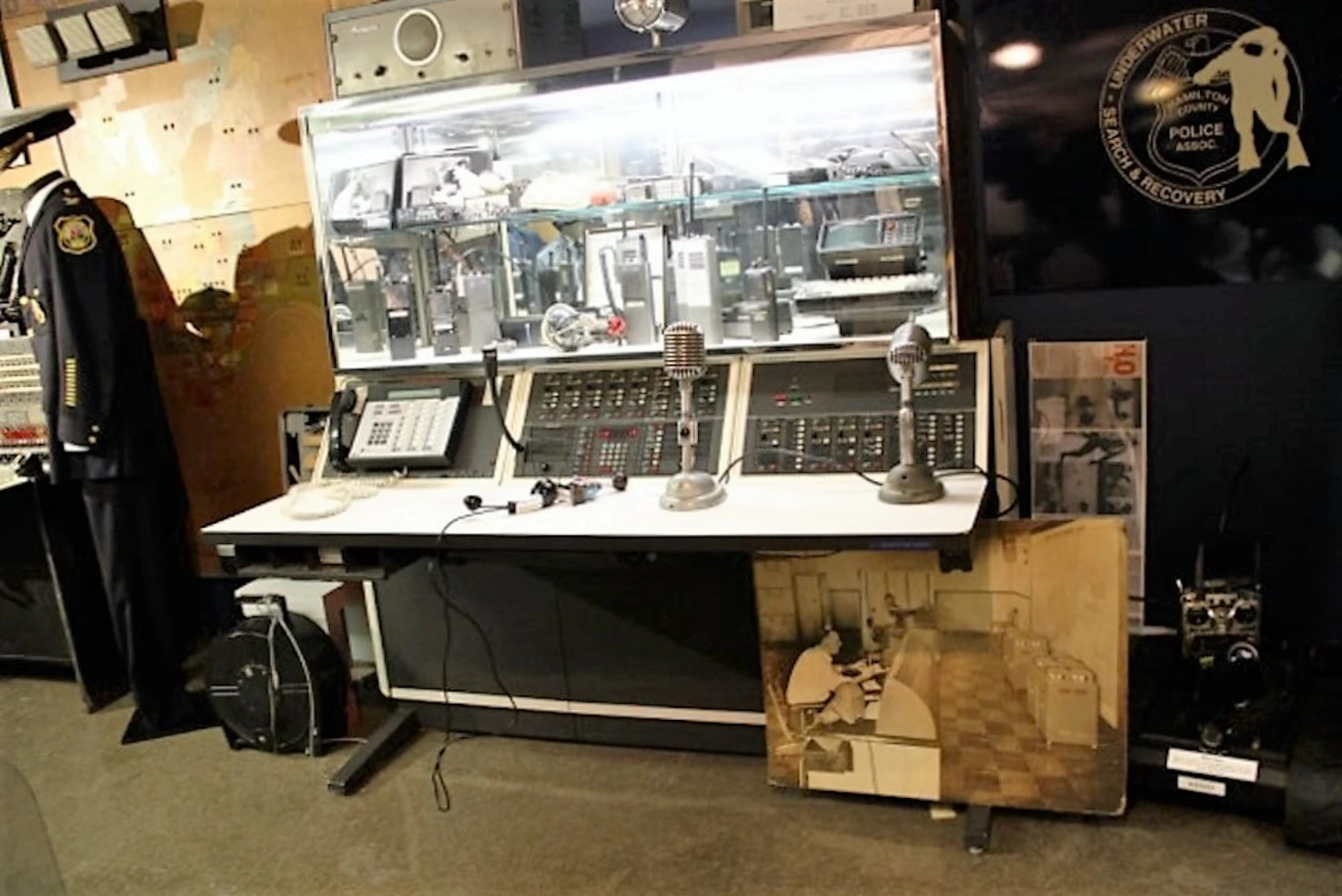 A vintage control console with microphones, buttons, and switches, flanked by communication equipment, and a black-and-white photo on the floor.