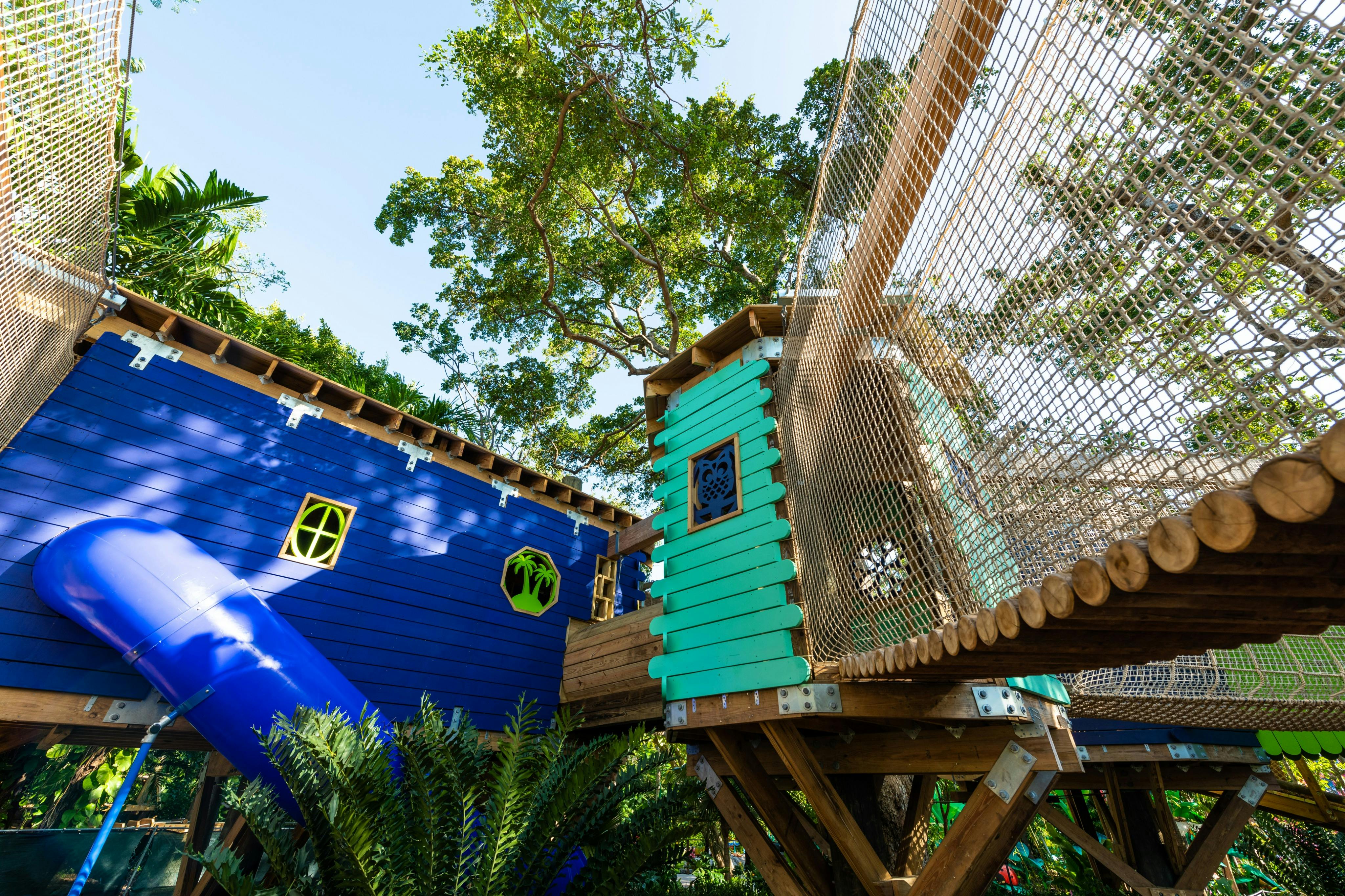 緑豊かな木々に囲まれた、ロープブリッジ付きの青とティールのツリーハウスを特徴とする木製の遊び場。