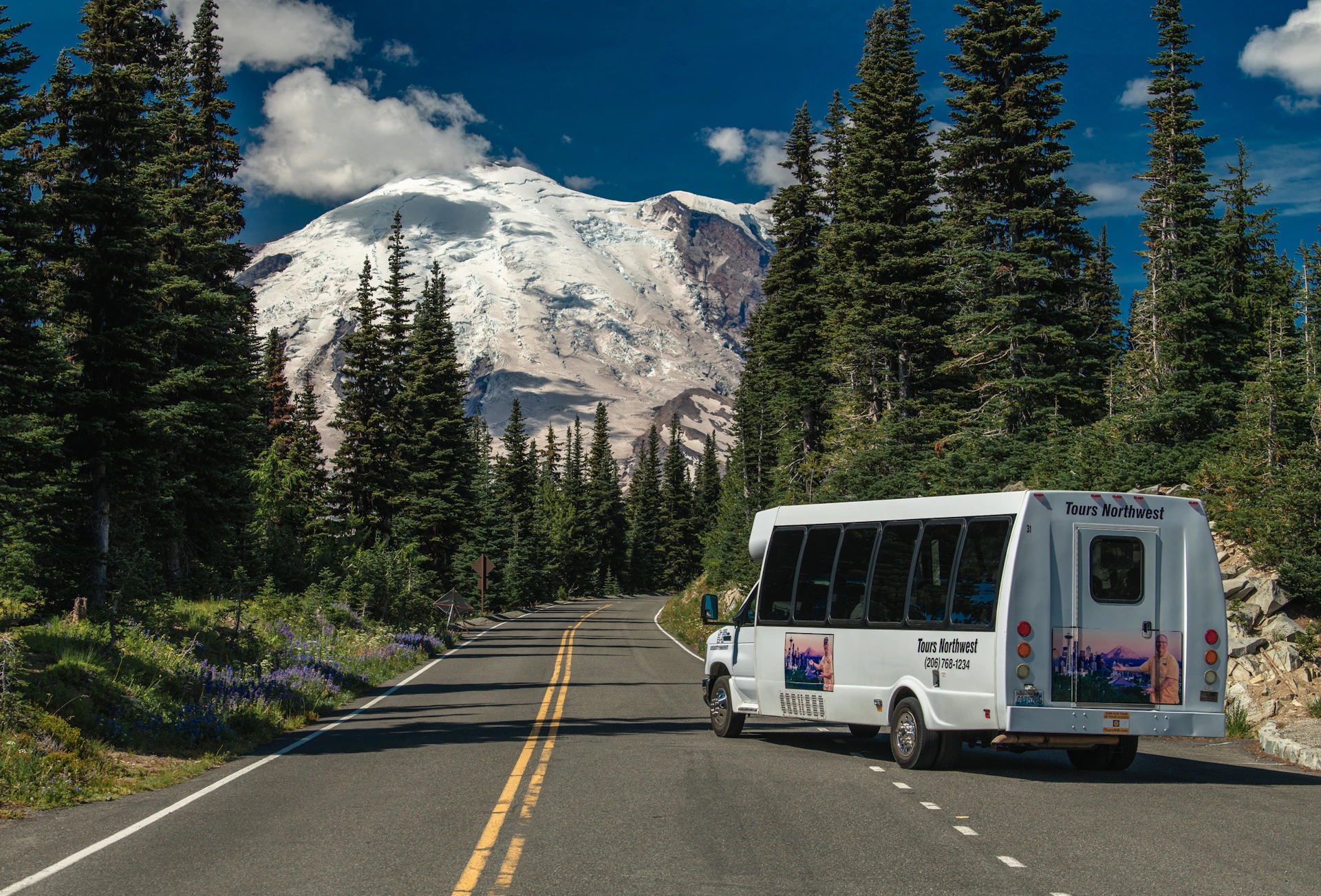 A tour bus on a forested mountain road with a snow-capped peak in the background, under a blue sky.