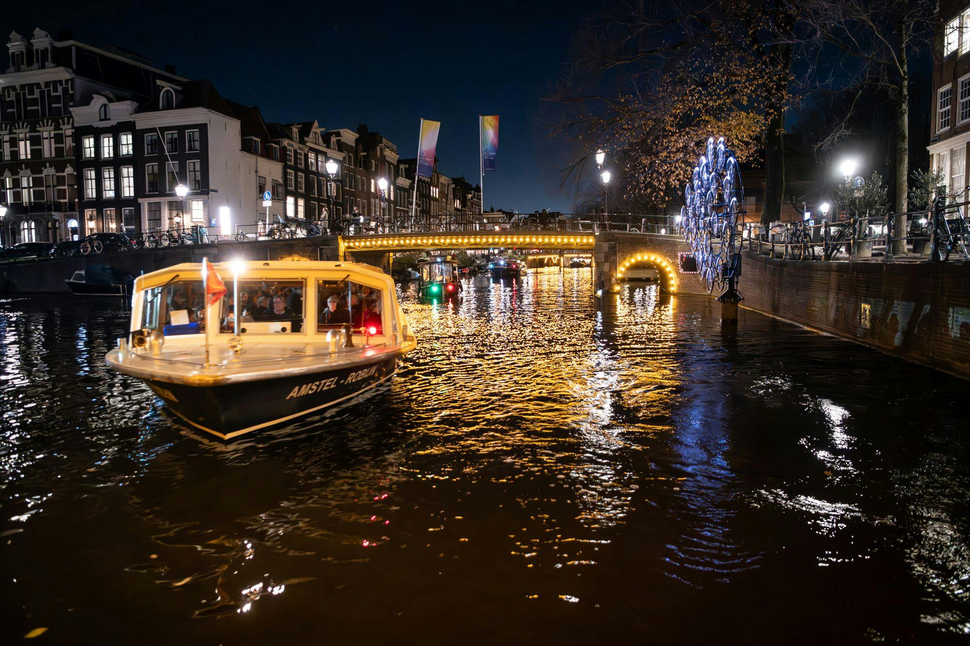 A canal at night with a boat, illuminated bridge, and festive lights. People are visible inside the boat.