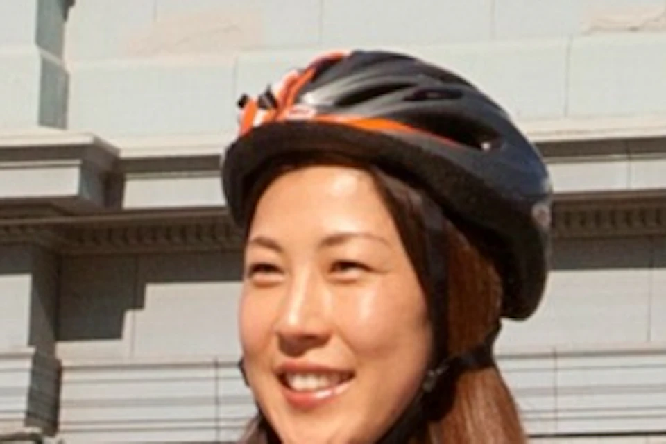 Two people in helmets riding bicycles in front of a large building with arched windows and a clock tower.