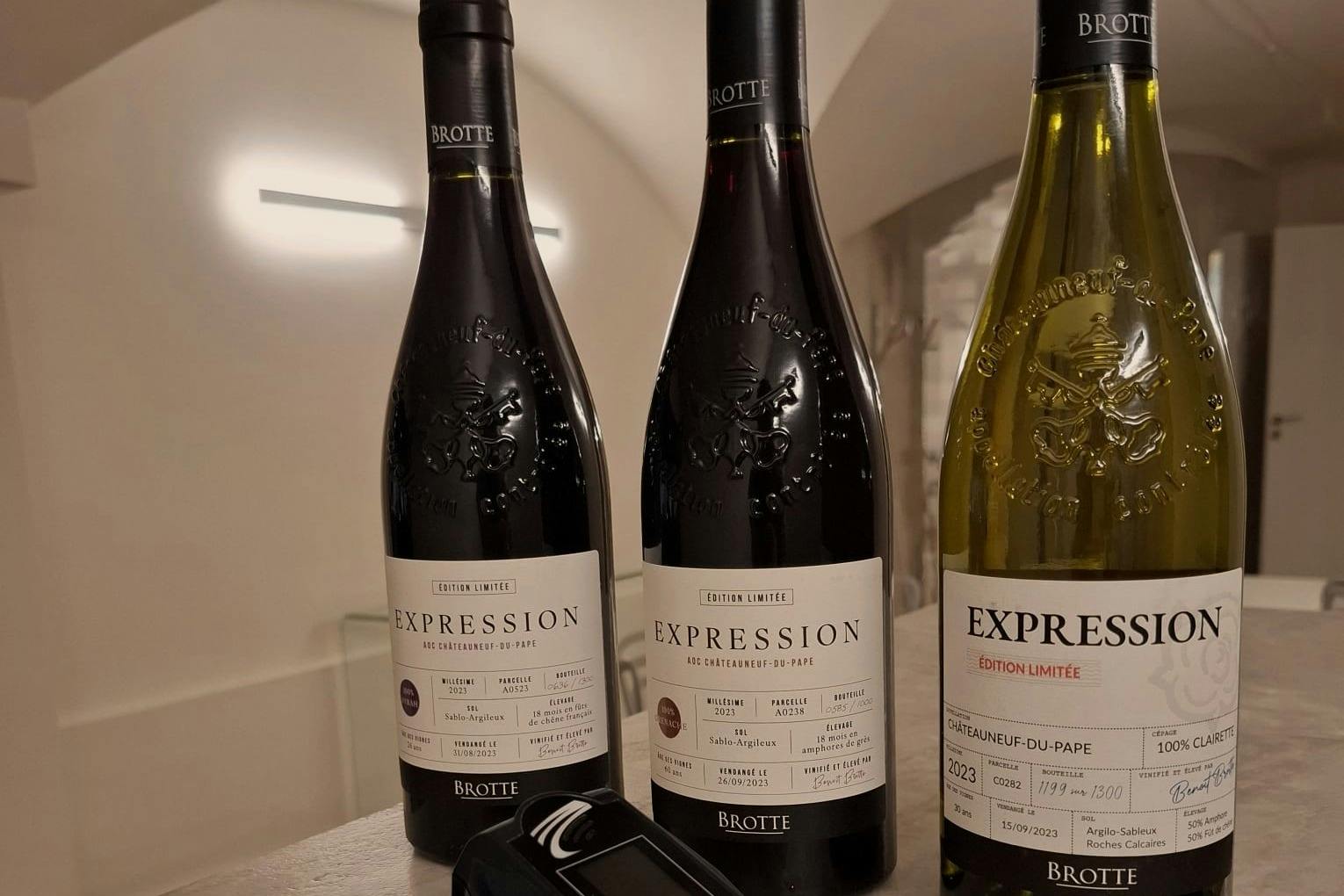 Τρία μπουκάλια κρασιού Brotte με την ένδειξη «Expression», με ένα κίτρινο και δύο σκούρα, τοποθετημένα σε ένα τραπέζι δίπλα σε μια ψηφιακή συσκευή.