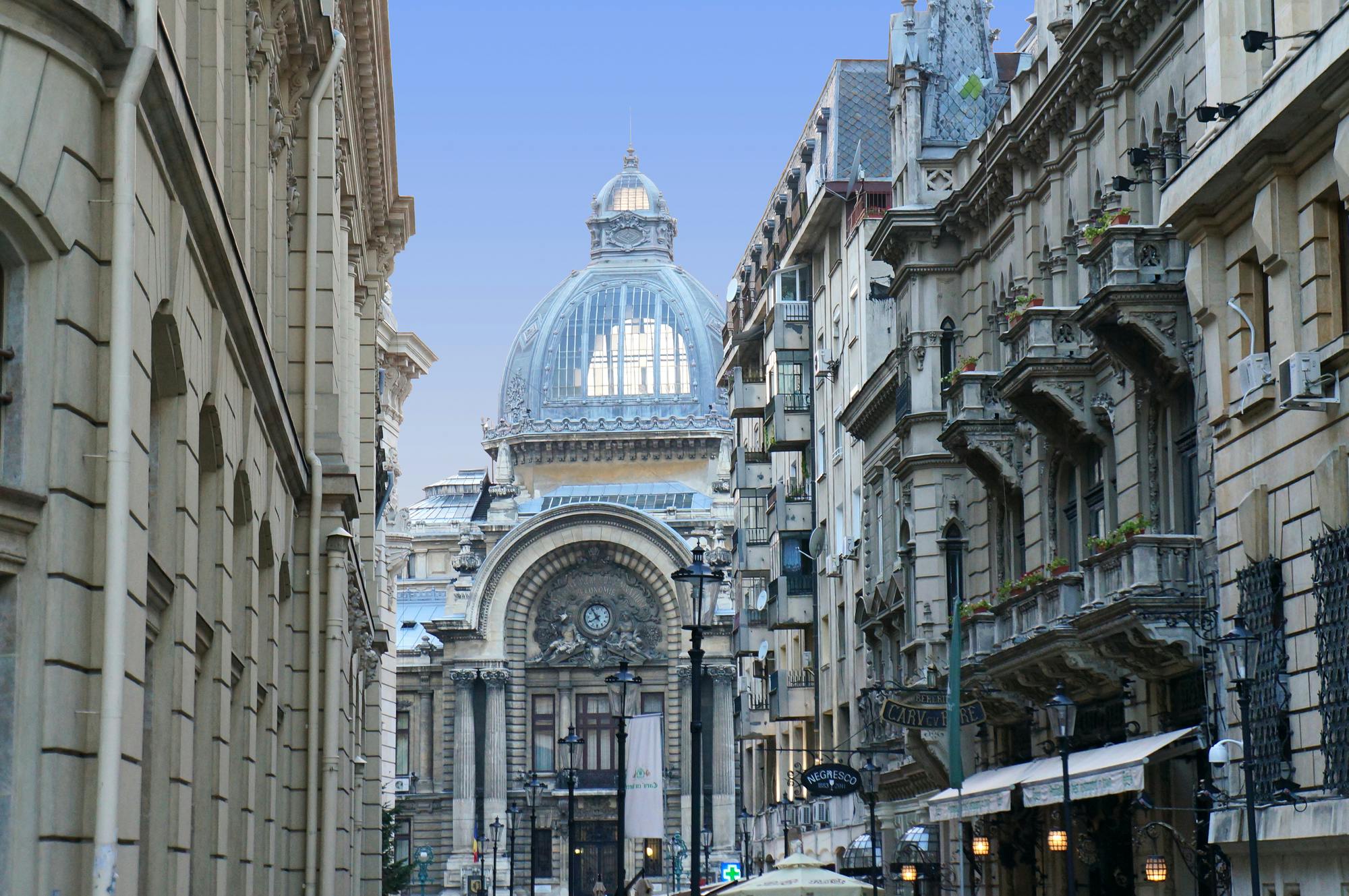 Narrow European street with historic, ornate buildings leading to a grand, domed structure. Cafe umbrellas in foreground.