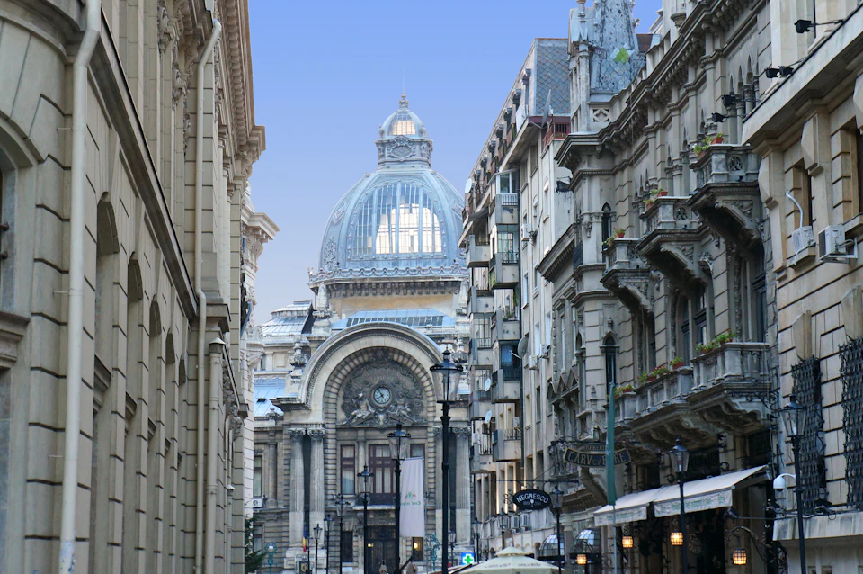 Narrow European street with historic, ornate buildings leading to a grand, domed structure. Cafe umbrellas in foreground.