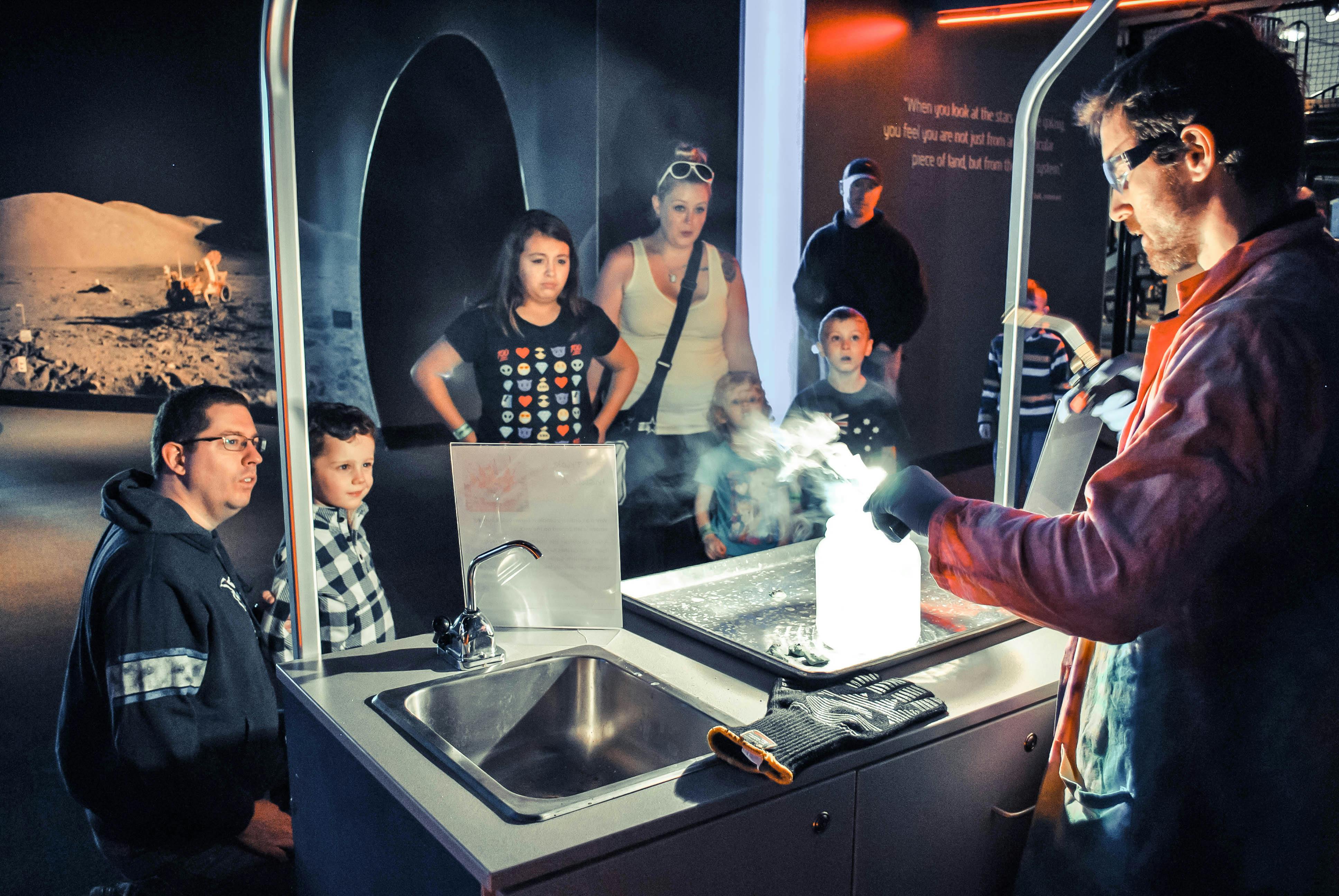 Um homem realiza uma experiência científica com produtos químicos, produzindo fumo, enquanto um grupo de crianças e adultos observa atentamente.