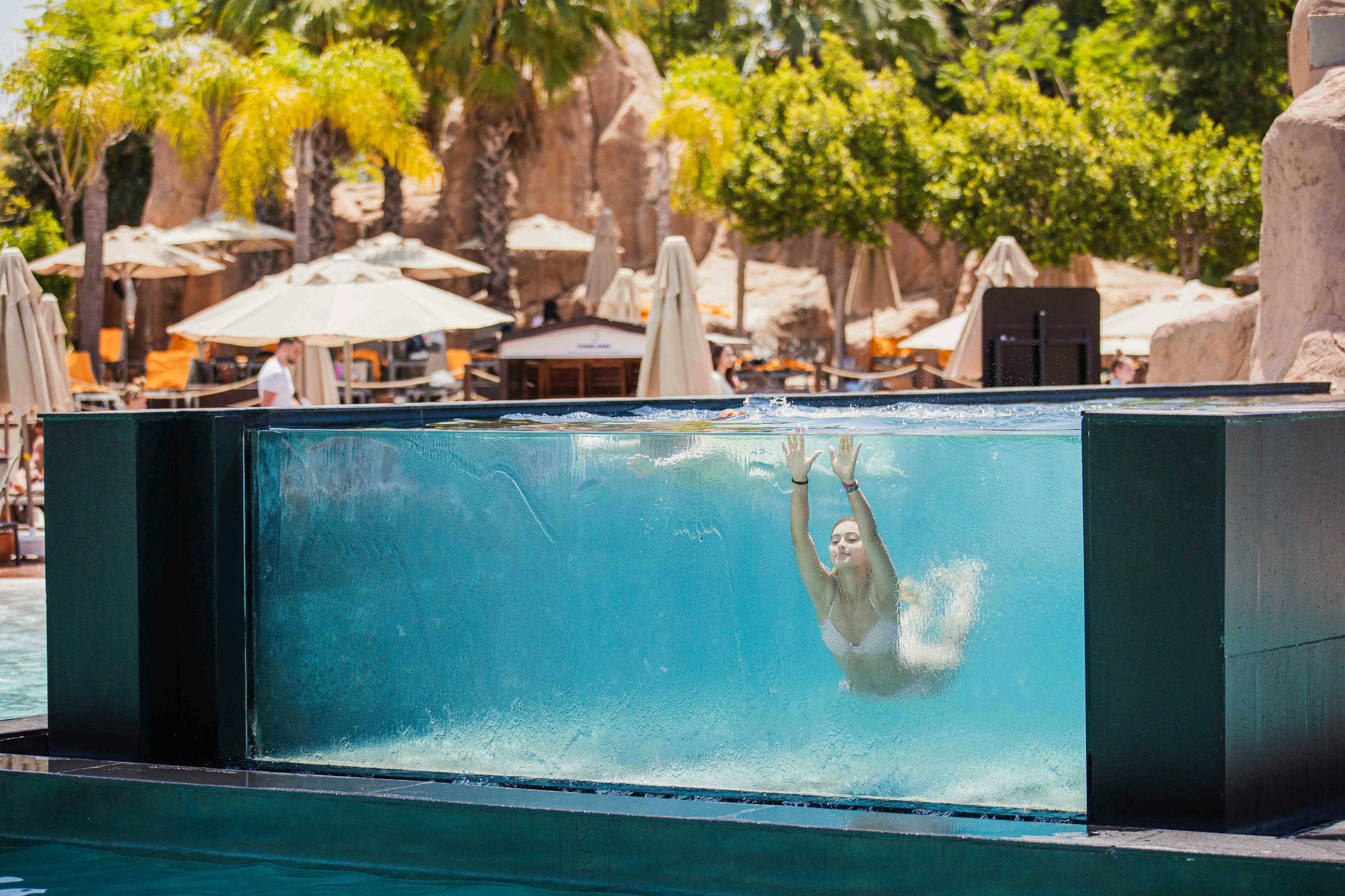 A woman swims underwater in a clear pool, with outdoor umbrellas and palm trees visible in the background.