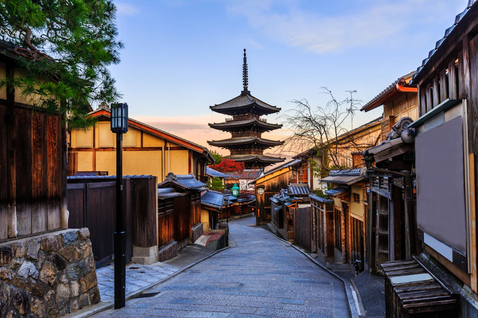 A traditional Japanese street with wooden buildings and a pagoda in the distance under a bright sky at sunset.