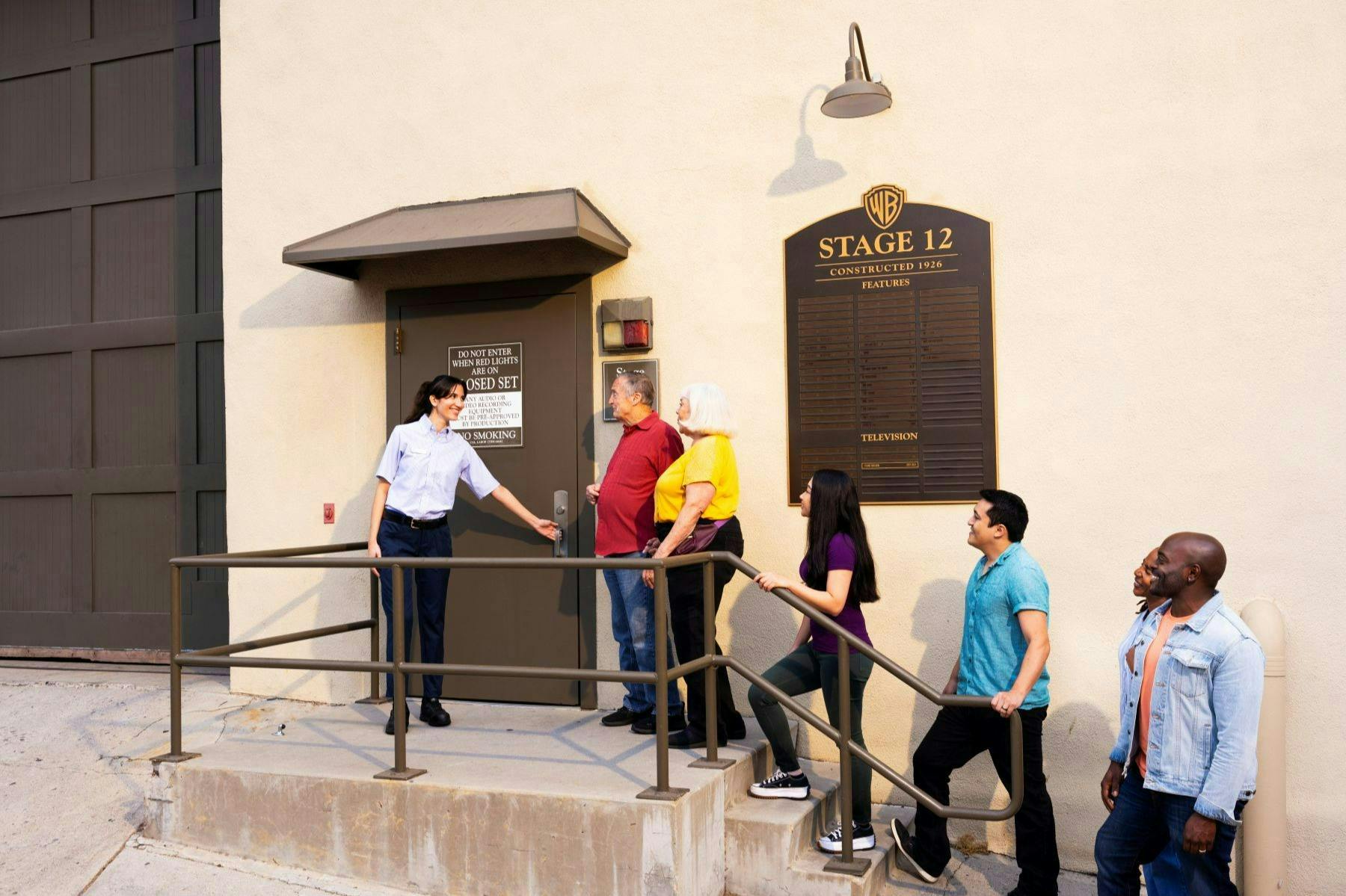 A group of people stands near a door labeled "Stage 12" with a guide in uniform holding the door open for them.