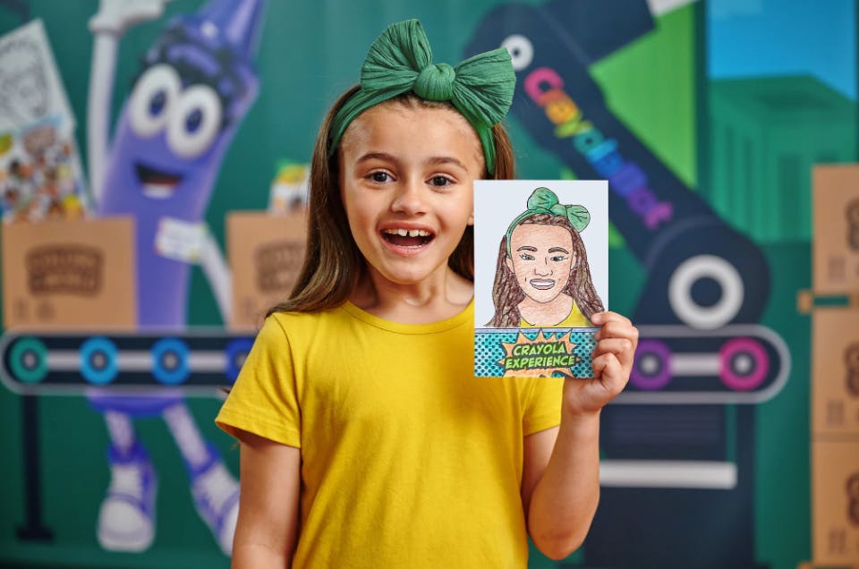 A young girl in a yellow shirt and green headband holds up a drawing of herself in front of a colorful background.
