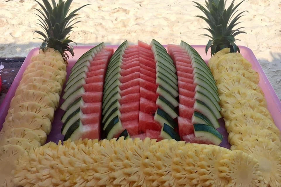 Sliced watermelons and pineapples arranged neatly on a table, with whole pineapples at the corners, displayed on a sandy background.