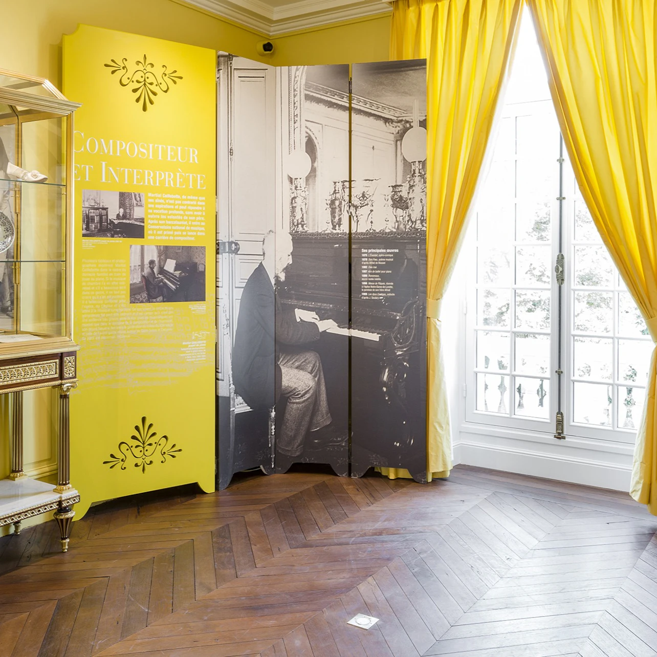 A museum display with a yellow panel, a historical photo partition, a glass cabinet, and tall yellow curtains over a window.