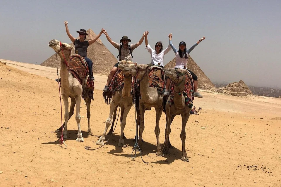 Four people riding camels in the desert, holding hands, with pyramids in the background.