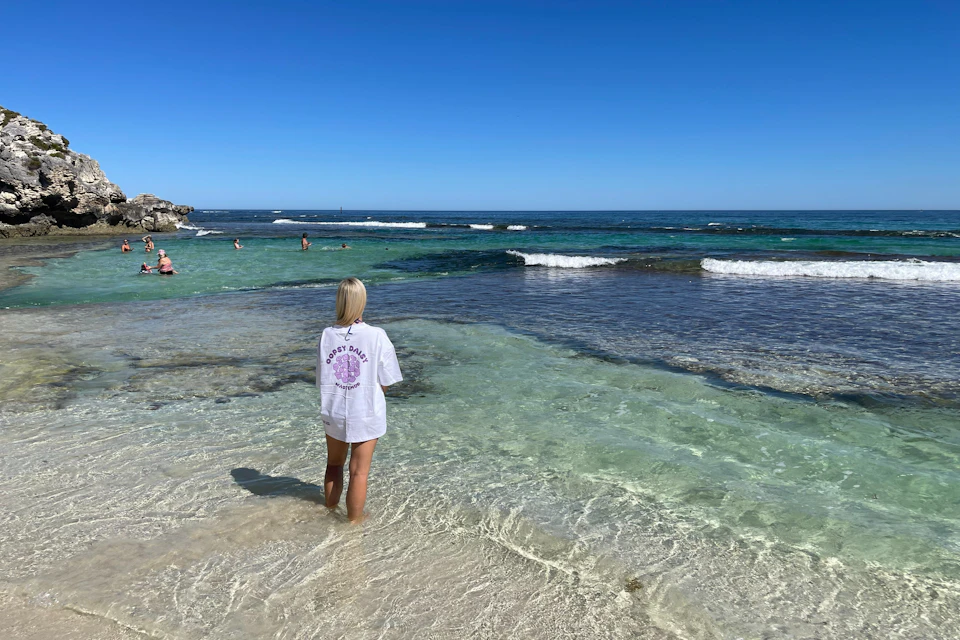 A person stands in shallow, clear water at a beach, facing the ocean under a clear blue sky.