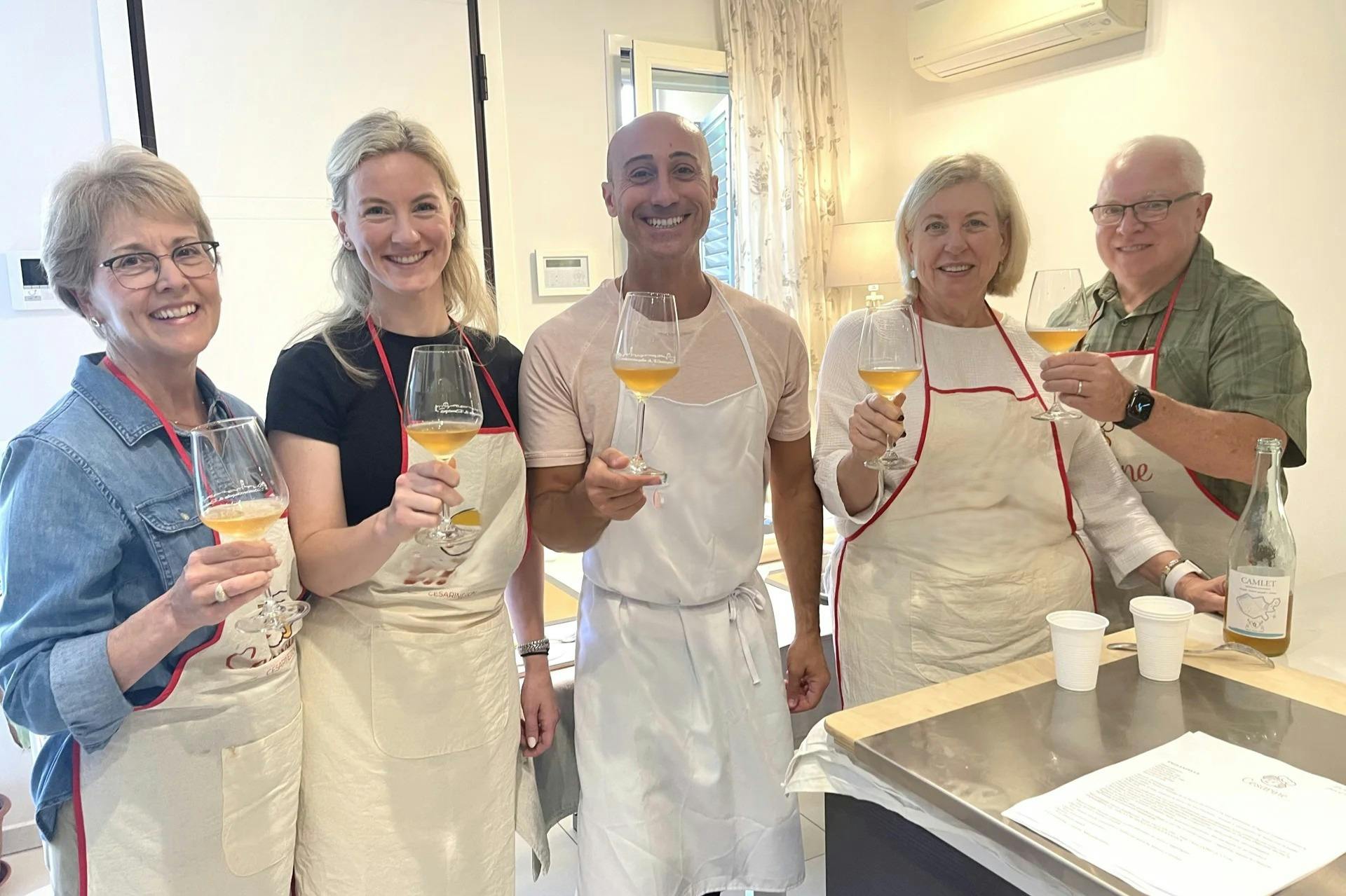 Three people wearing aprons toast with glasses of orange wine in a brightly lit room.