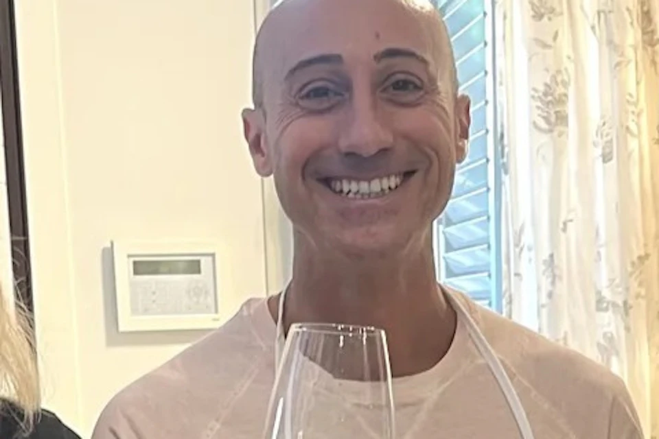 Three people wearing aprons toast with glasses of orange wine in a brightly lit room.