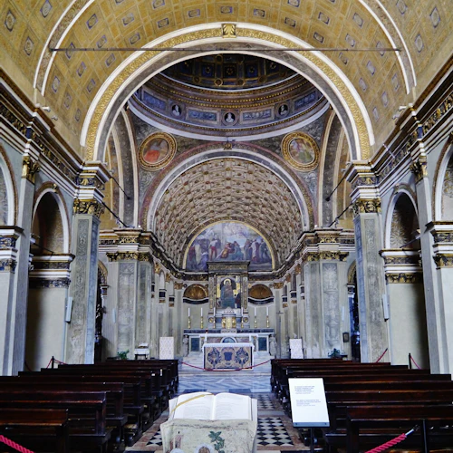 Interiör av en stor kyrka med utsmyckade valv, fresker, ett altare och flera rader av träbänkar.