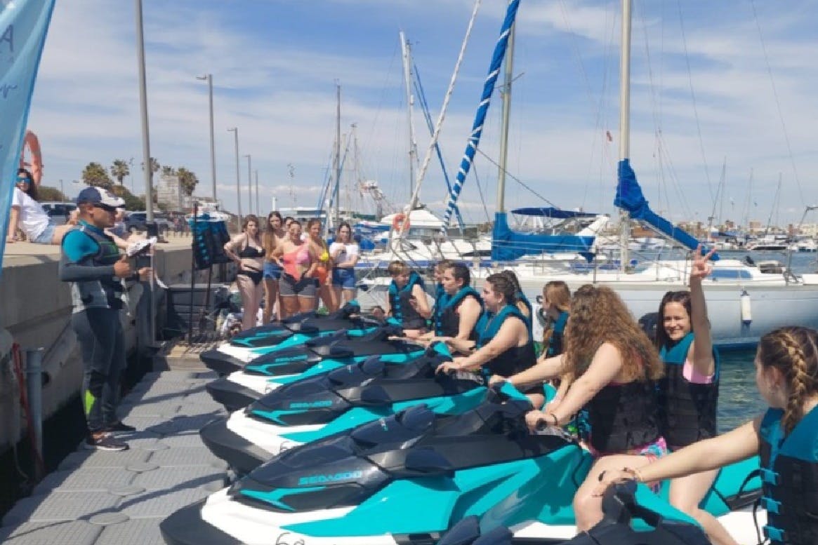 Skupina lidí v záchranných vestách sedí na vodních lyžích v přístavu, zatímco jiní v plavkách stojí nedaleko a v pozadí jsou vidět lodě.