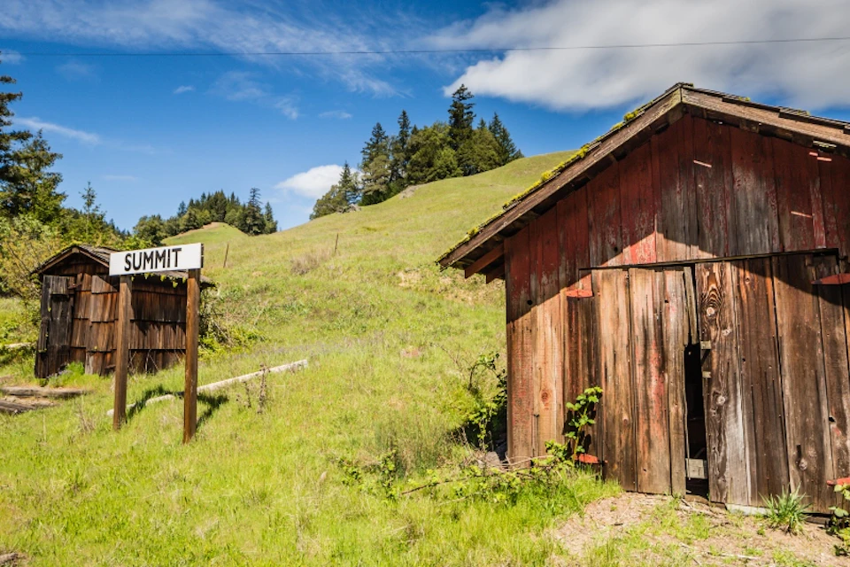 A wooden shed stands next to a grassy hill under a blue sky. A sign with "Summit" is partially visible on the left.