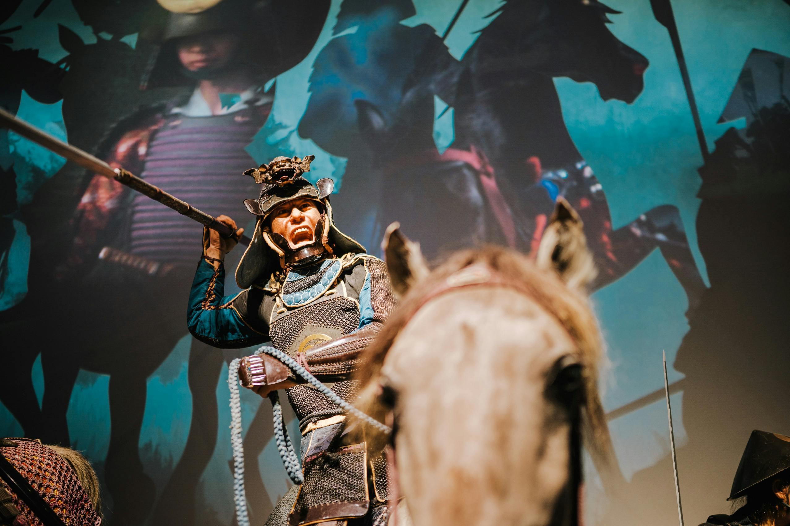 A person dressed as a samurai warrior holding a spear, with a dramatic backdrop featuring horse riders in shadow.