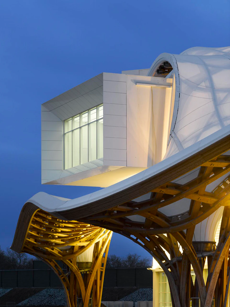 Modern building with a large, illuminated white box structure extending from a curved roof made of wood and fabric, against a twilight sky.