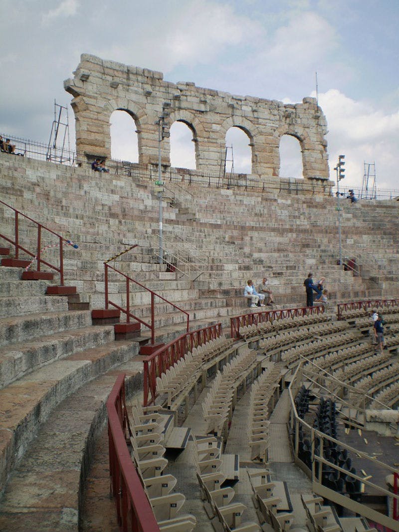 古老的石头圆形剧场，空荡荡的座位，三个大拱门，几个人走在台阶上，坐在台阶上。