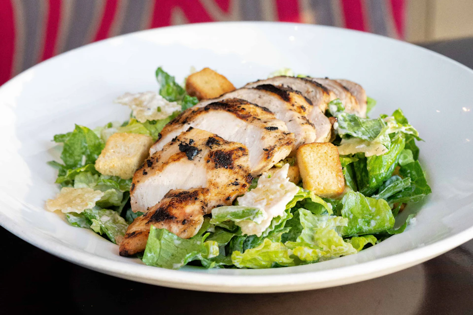 Grilled chicken slices on a bed of Caesar salad with croutons in a white bowl.