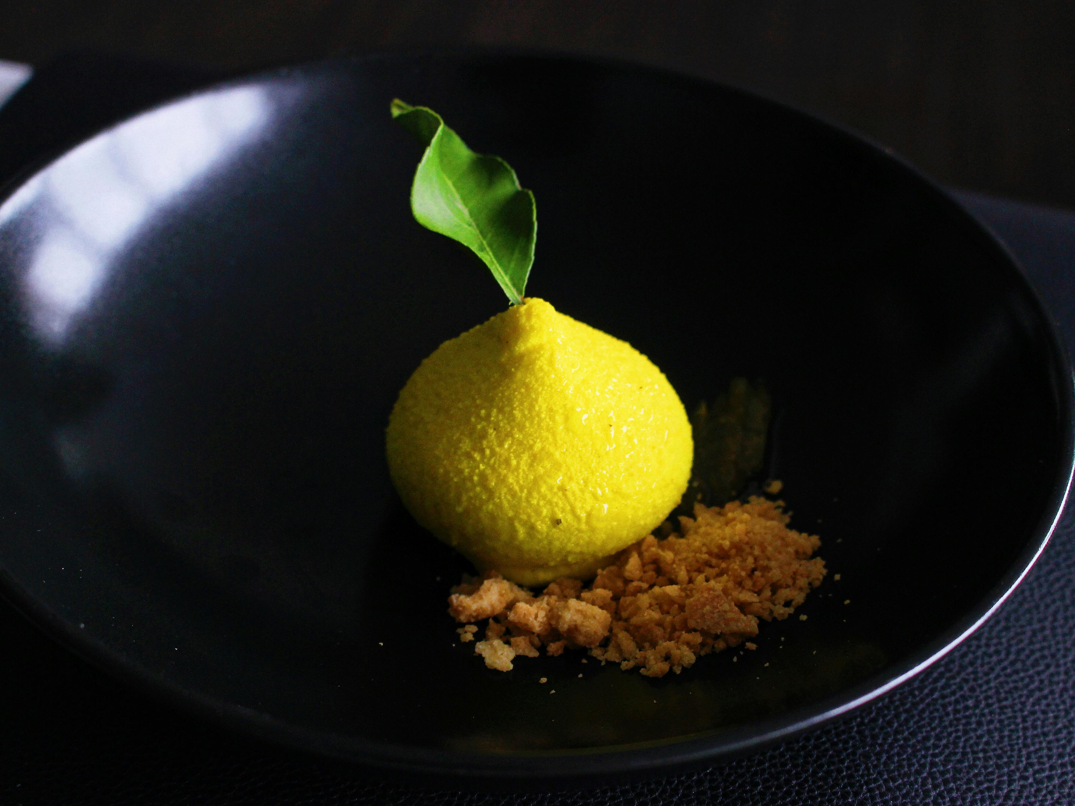 黄色圆顶形甜点，配有绿叶装饰，放在黑色盘子上，旁边有碎顶。