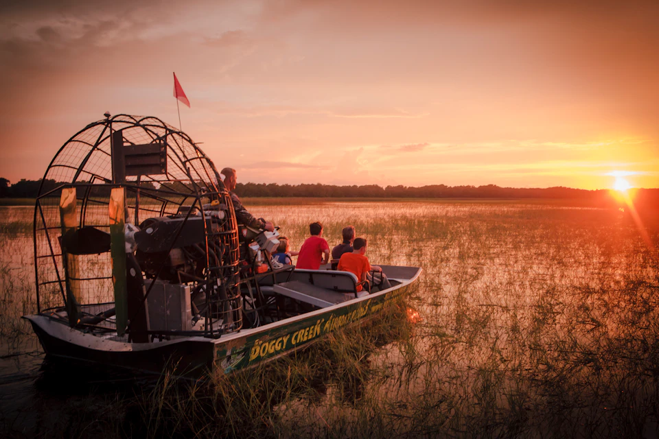 People on an airboat ride through a marsh at sunset, with a person standing in the front and others seated, viewing the landscape.