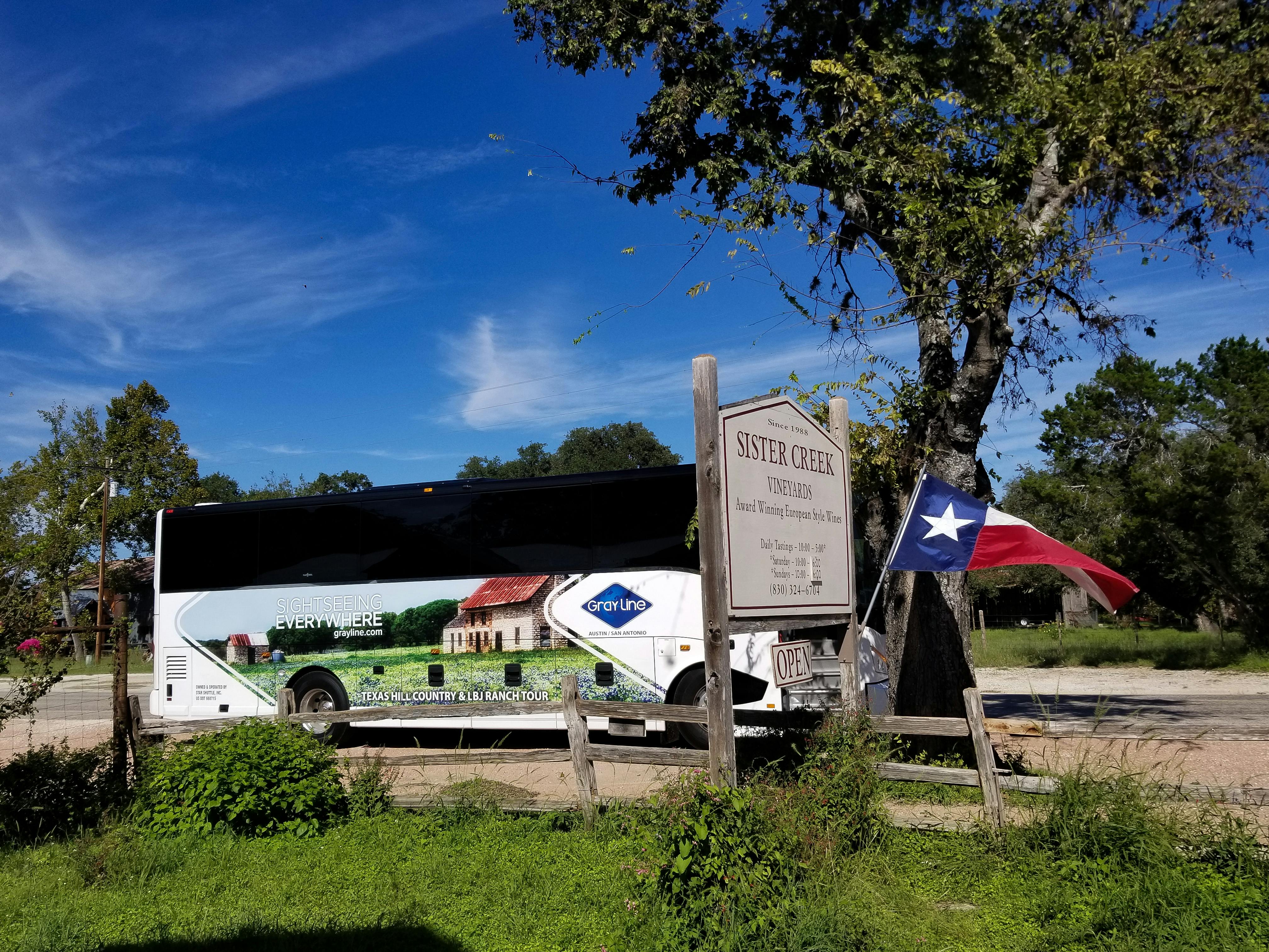 U dřevěného plotu je zaparkovaný turistický autobus s cedulí na vinice Sister Creek s texaskou vlajkou a velkým stromem v pozadí.