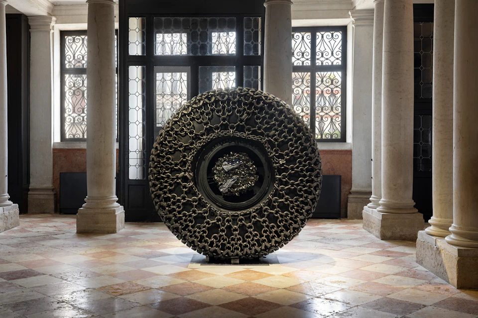 A circular sculptural artwork made of interwoven chains displayed in a room with ornate windows and marble columns.