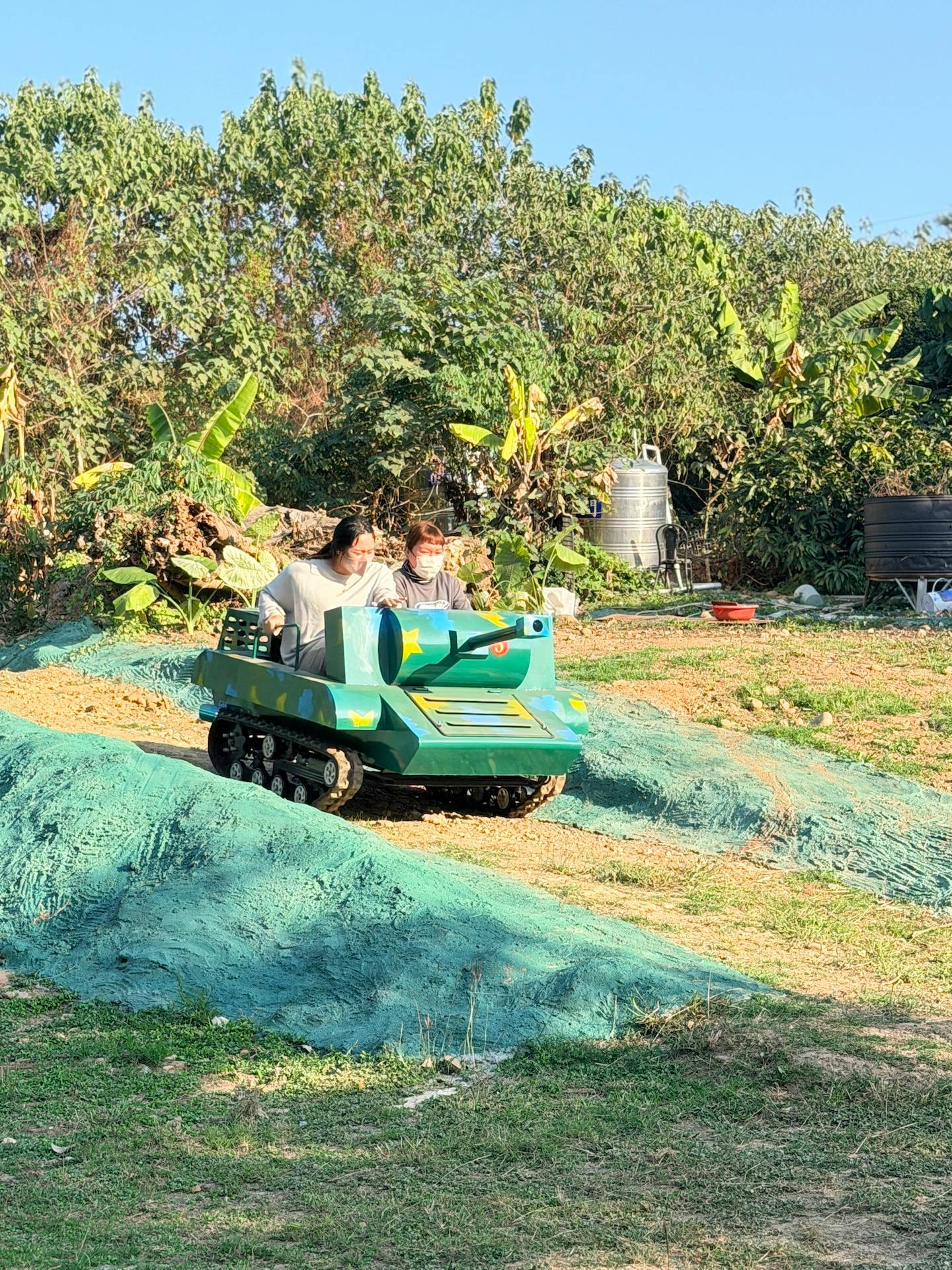 Two people riding a small green tank-shaped vehicle on a grassy and hilly terrain with trees in the background.