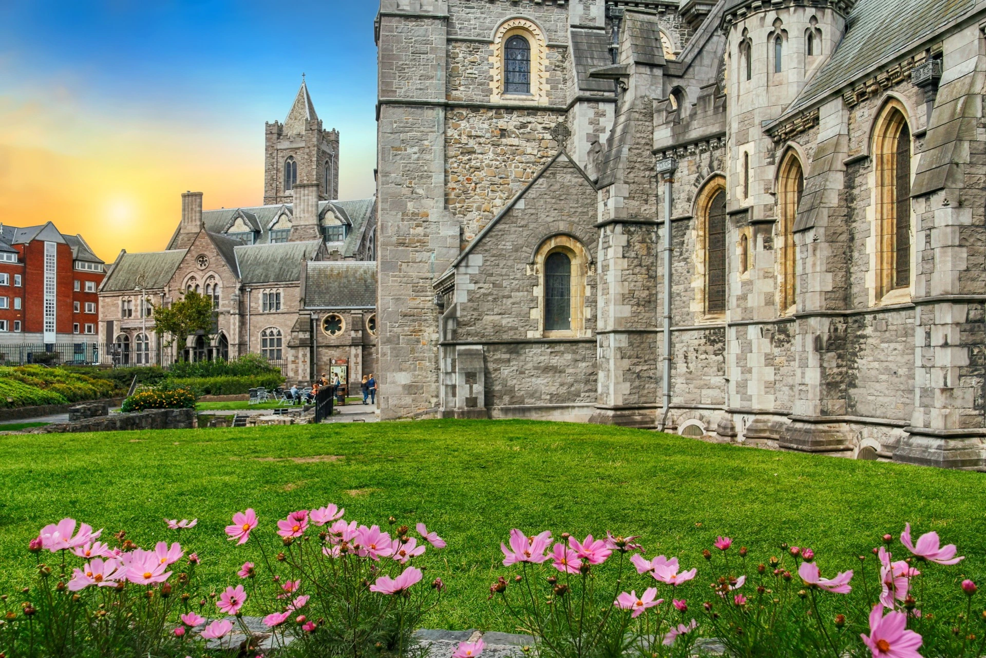 Historic stone church with Gothic architecture, flowering pink plants in the foreground, well-kept lawn, and a blue sky at sunset.