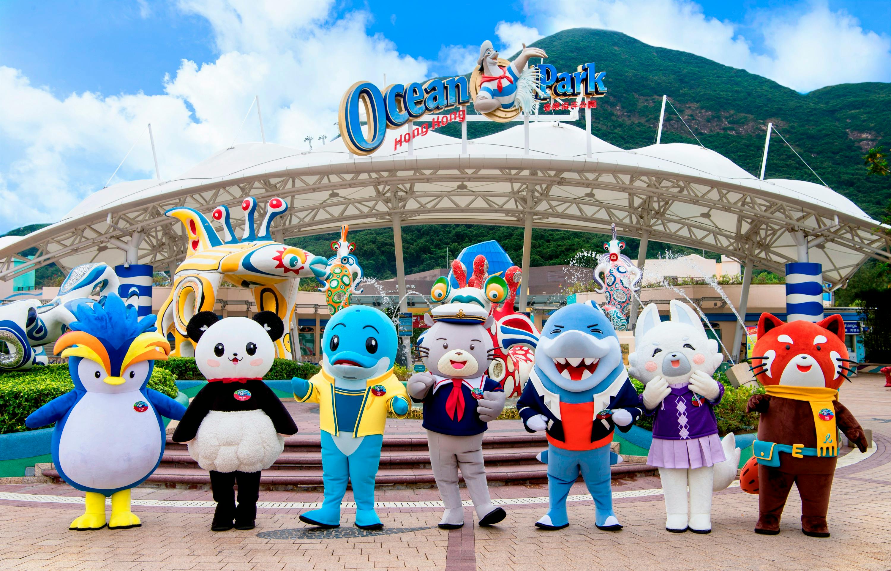 Mascots in colorful costumes stand in front of the Ocean Park Hong Kong entrance with a mountain in the background.