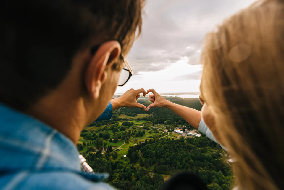Two people form a heart shape with their hands, overlooking a vast green landscape under a cloudy sky.