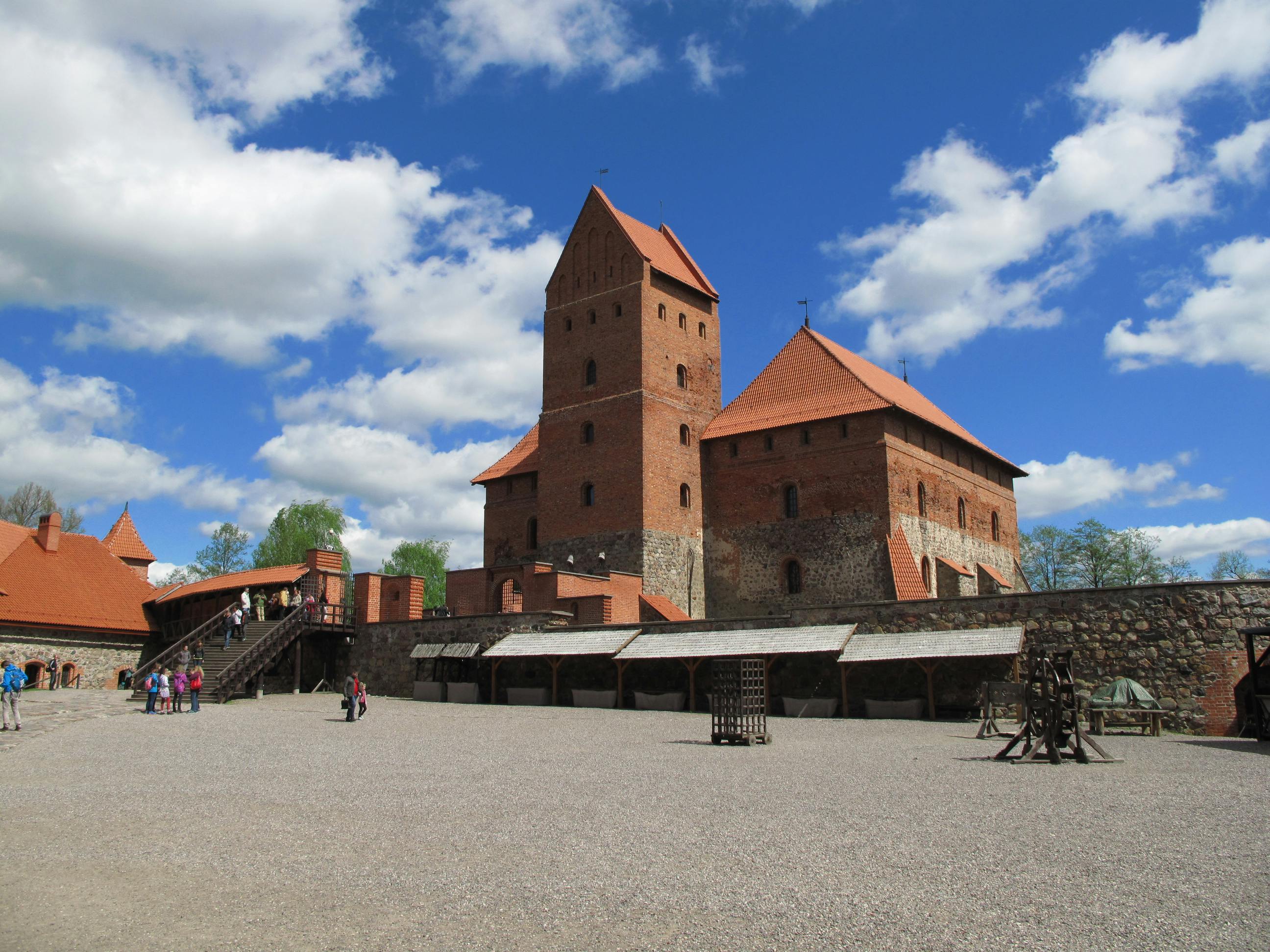 Ένα μεσαιωνικό κάστρο με κόκκινους τούβλινους πύργους και ένα πέτρινο τείχος, που περιβάλλεται από ανθρώπους και κατασκευές σε μια ηλιόλουστη μέρα με καθαρό ουρανό.