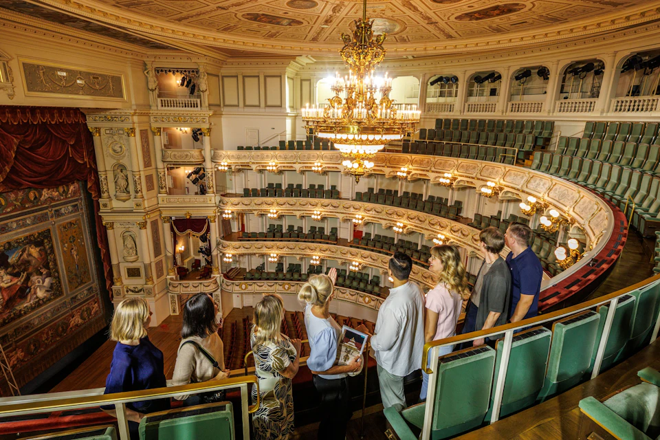 A group of people tours an ornate theater with tiered balconies, decorative details, and a grand chandelier.