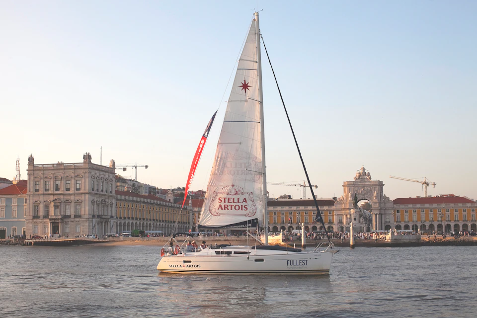 Sailboat with "Stella Artois" branding in a bay near a historic building under a clear sky.