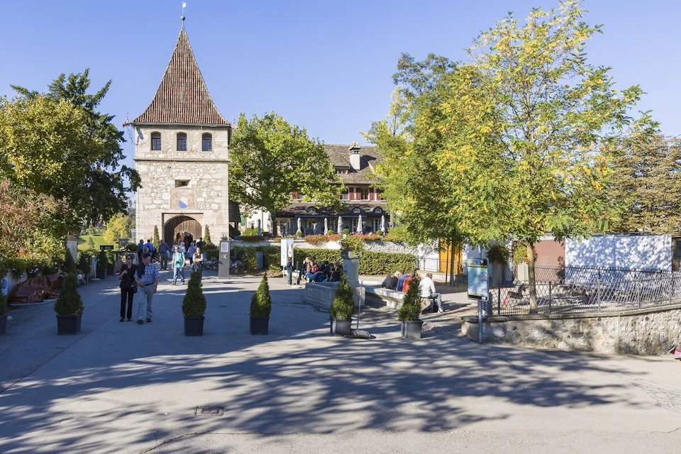 A stone building with a pointed roof stands in a courtyard, surrounded by trees and people enjoying the sunny day.