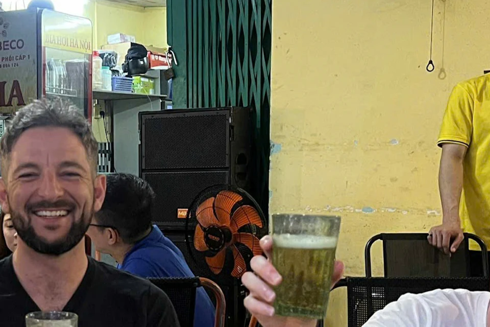 Three men sitting at a table in a casual restaurant, smiling and holding up glasses. Other diners and a standing waiter in the background.