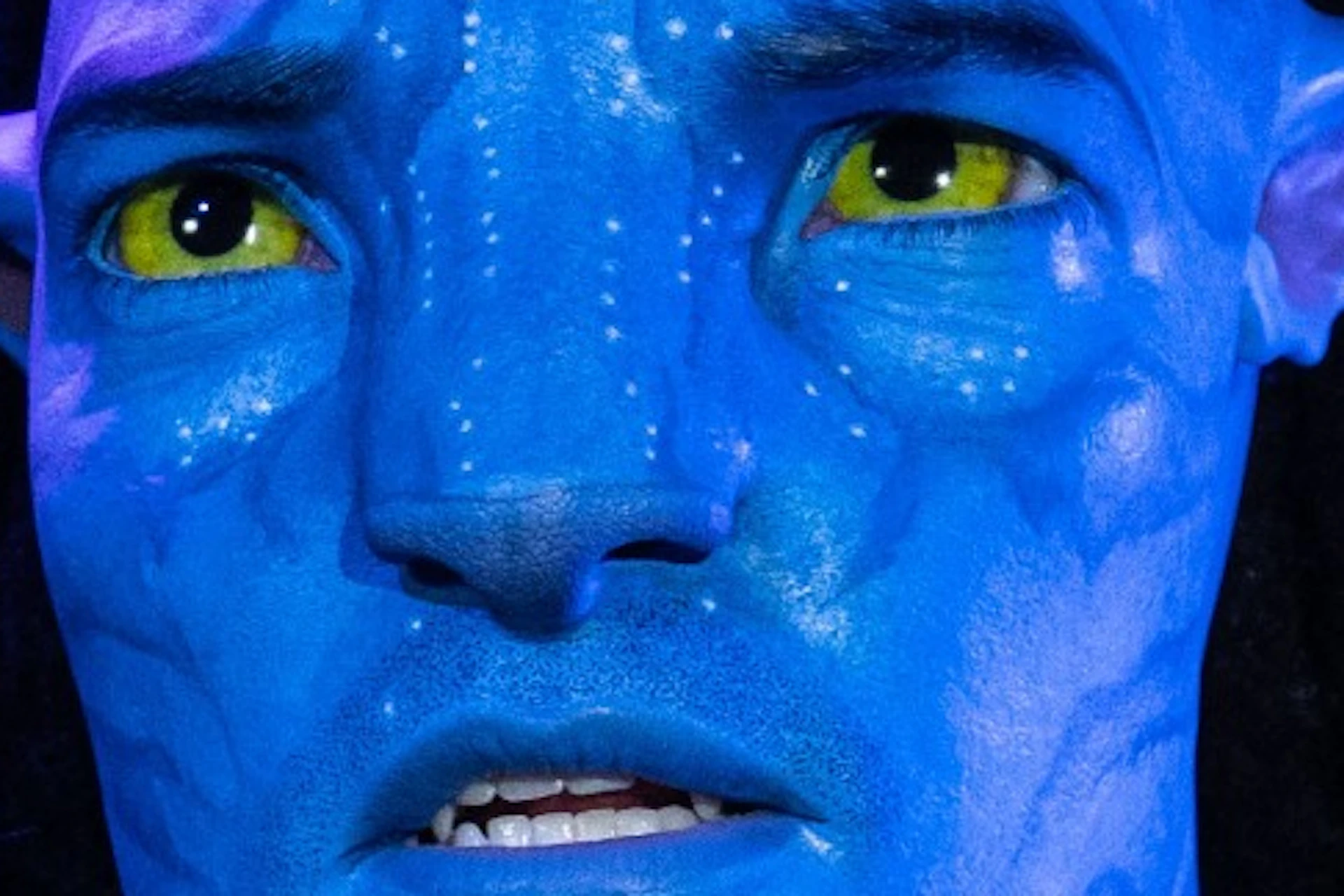 A blue-skinned humanoid figure with glowing yellow eyes and braided hair, wearing tribal and tactical attire, in a dimly lit environment.