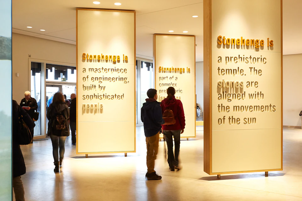 People walking through a museum exhibit with illuminated panels containing text about Stonehenge.